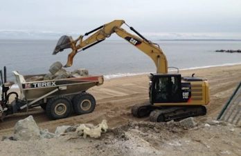 A yellow excavator loading sand into a white Terex dump truck on a beach.