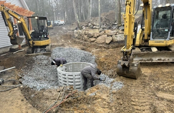 Two excavators positioned around workers installing a cylindrical concrete drainage well.