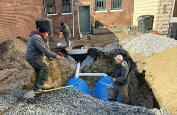 Workers installing blue plastic stormwater drainage chambers in an excavated trench.
