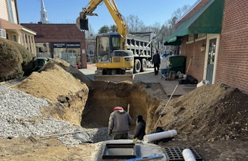 A yellow excavator parked along an excavated trench in an alleyway.