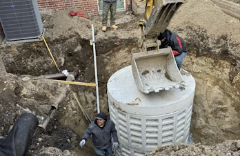 Workers and an excavator positioning a large slotted concrete tank in an excavation pit.